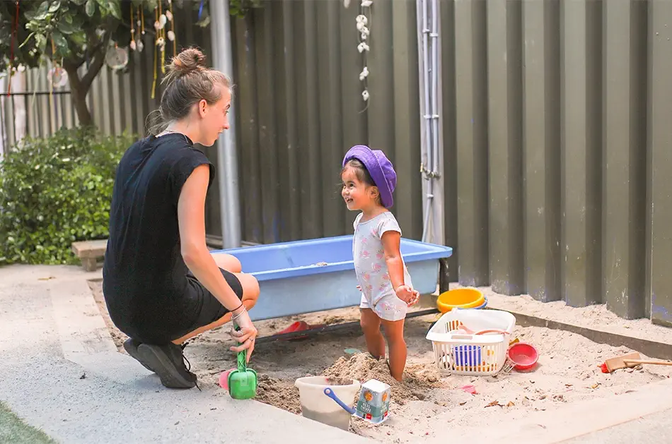 A teacher is kneeling down beside a child playing in the sandpit.